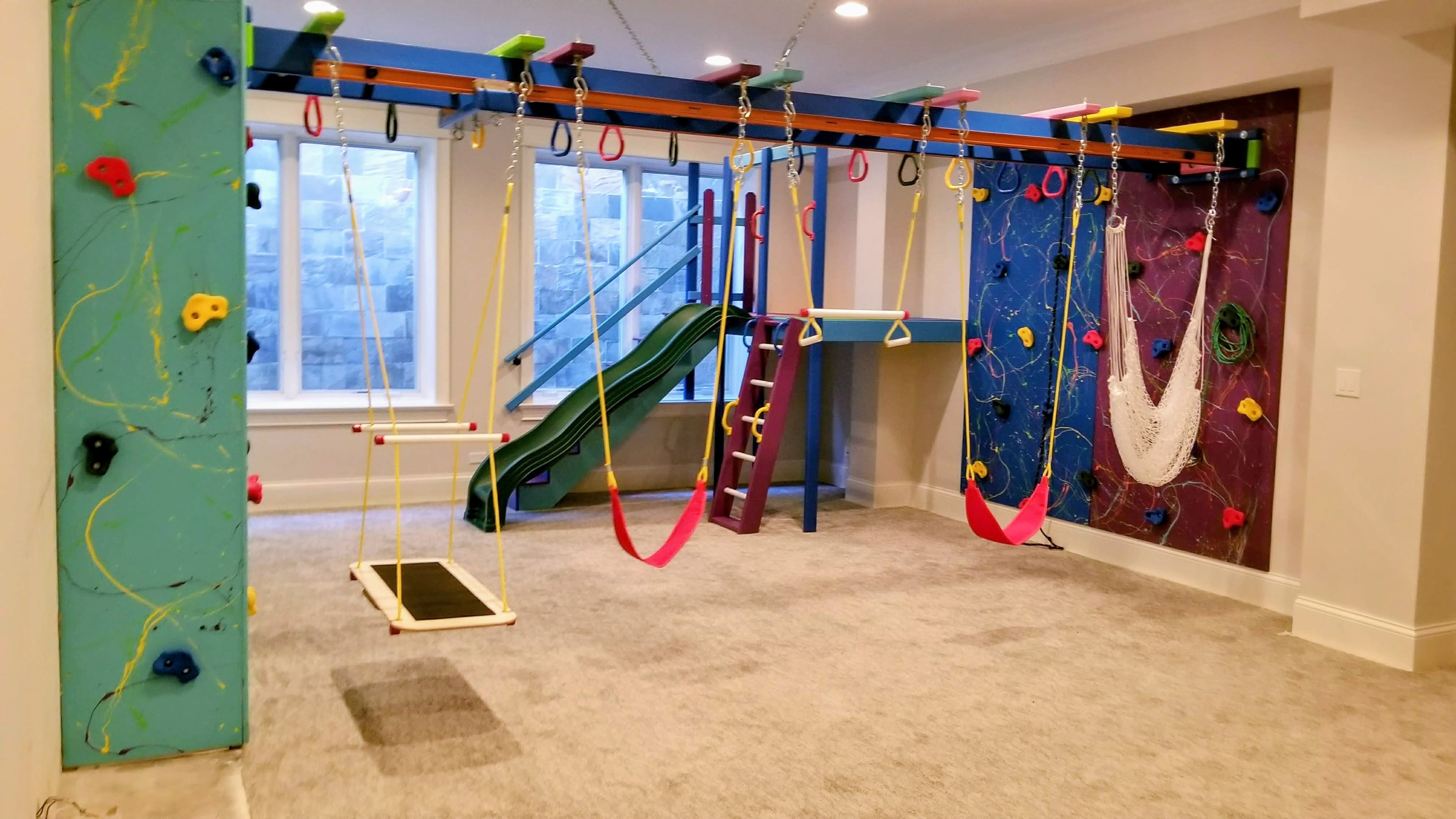Bright residential basement gym with swings, green slide, and climbing walls