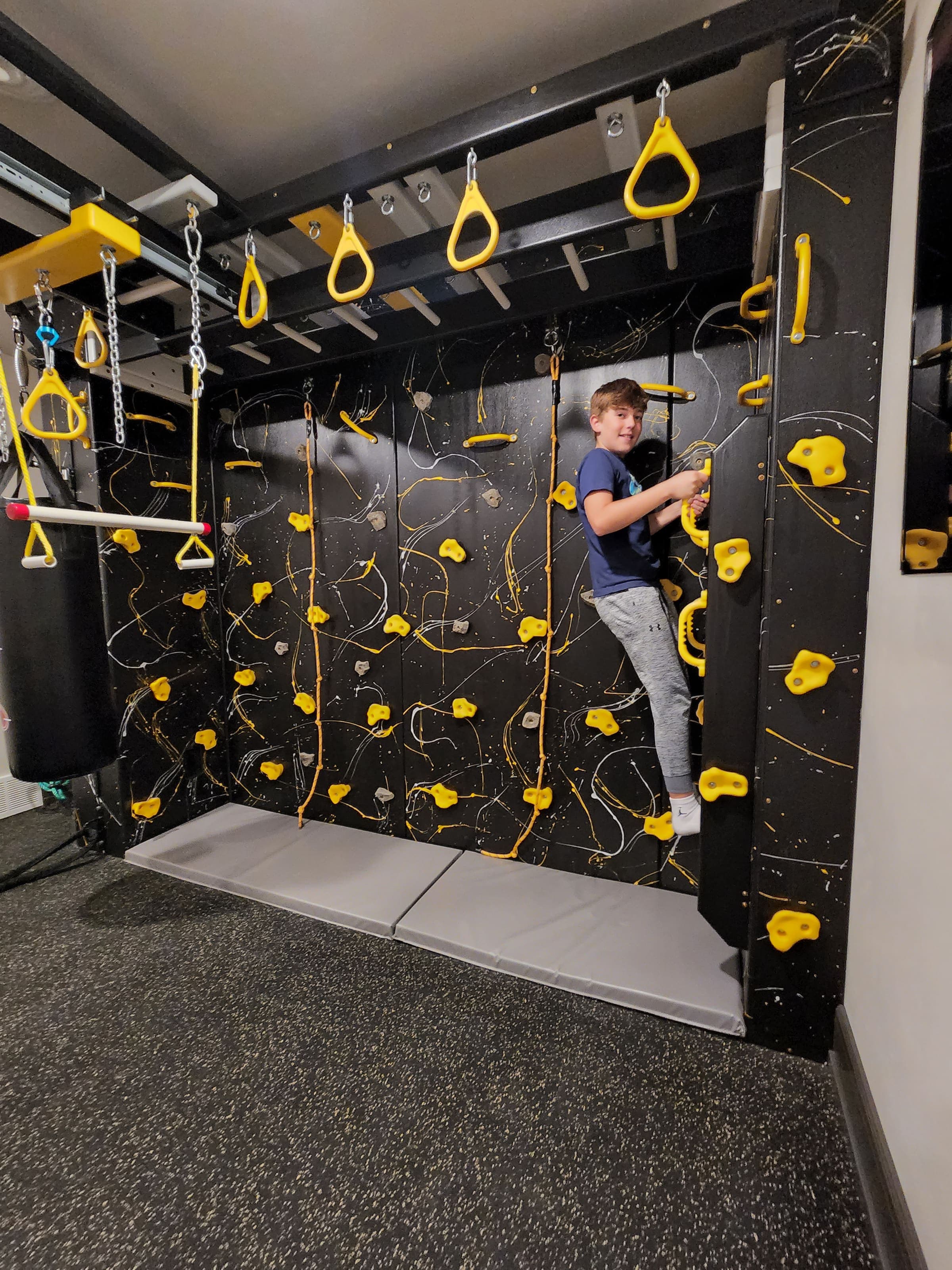 Boy climbing a black splatter wall with yellow holds under a ring beam in a home sensory gym