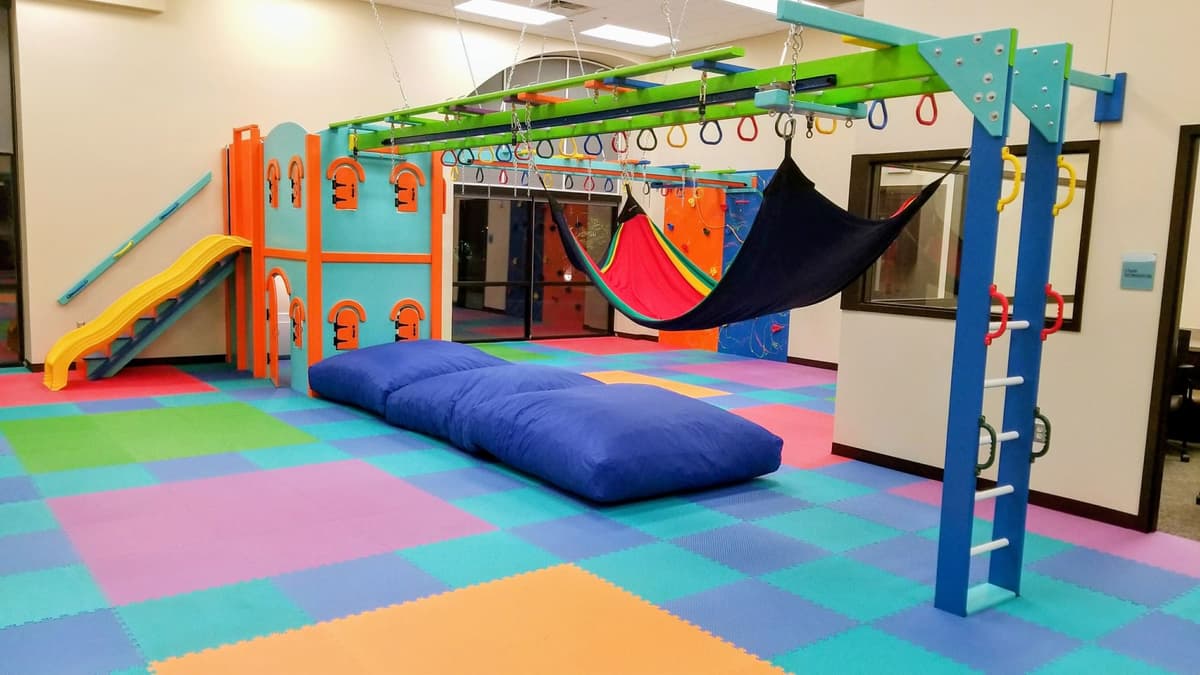 Pediatric OT clinic sensory gym with castle playhouse, yellow slide, hammock swings, and crash pillows