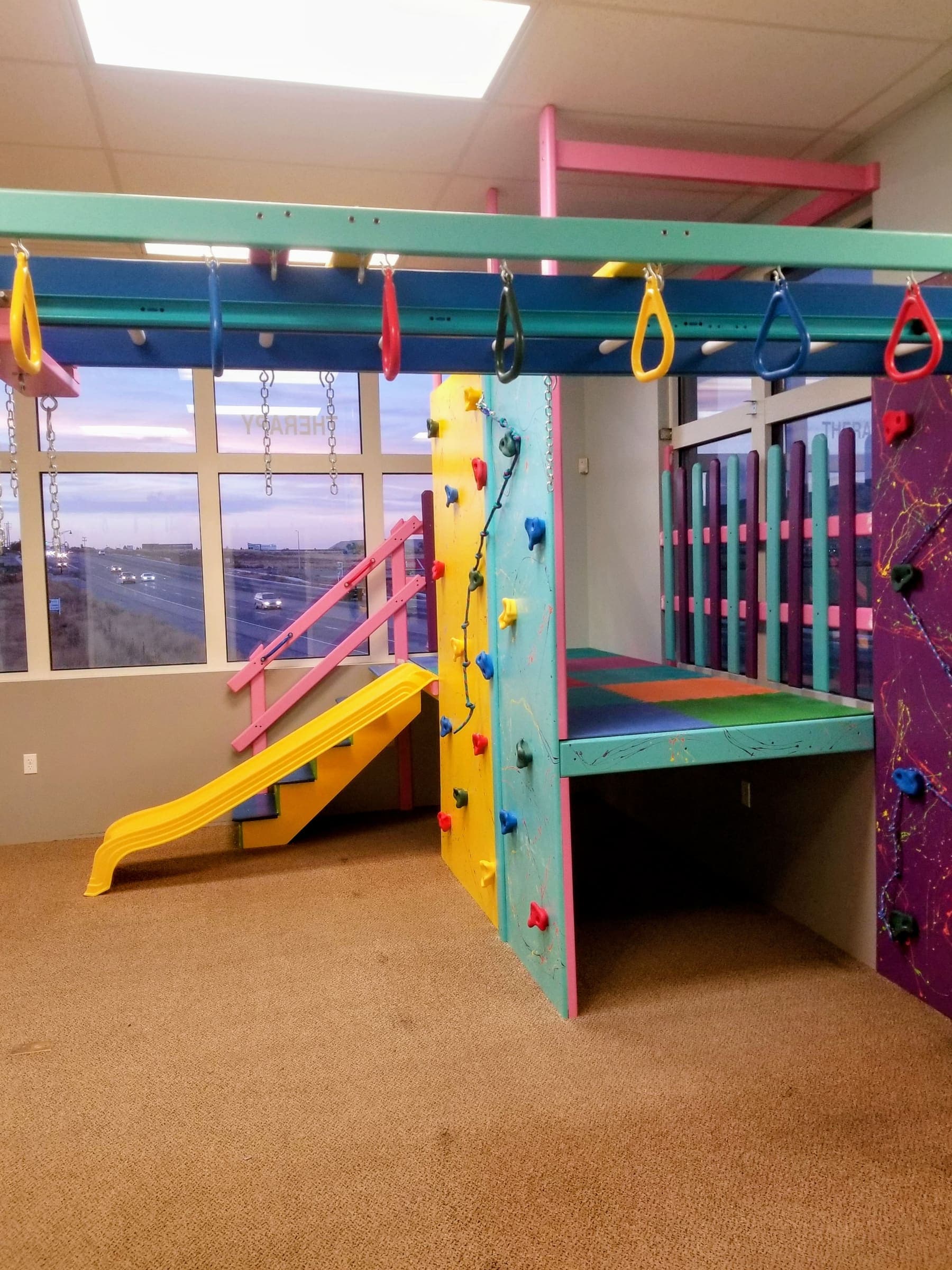 Sensory gym with yellow slide, pink stair rail, and splatter climbing wall beside large windows