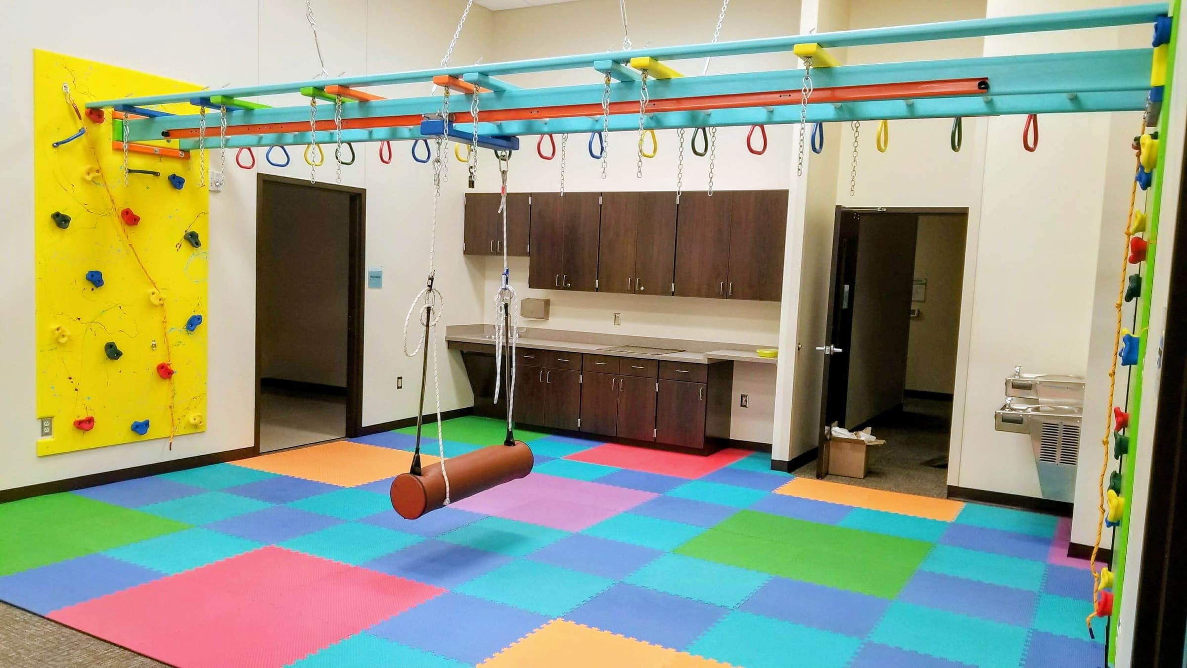 Wall-to-wall overhead ring beam with bolster swing and yellow climbing wall for vestibular input
