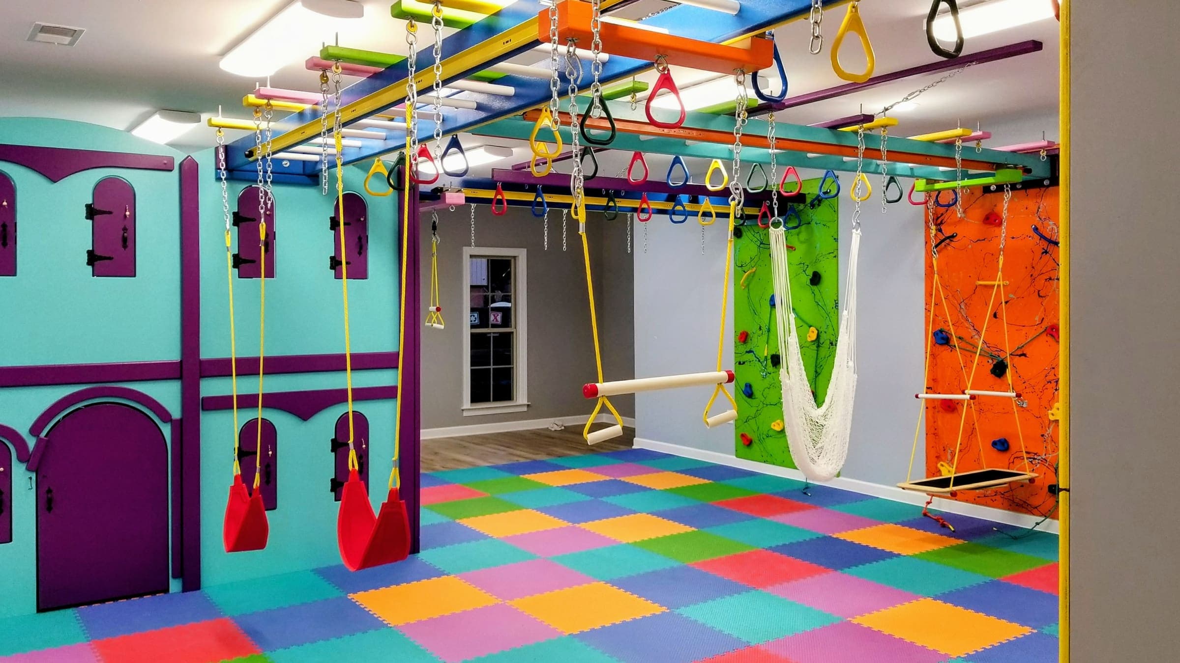 Sensory gym with castle playhouse, red swings, rope hammock, trapeze bar, and climbing walls on foam mats