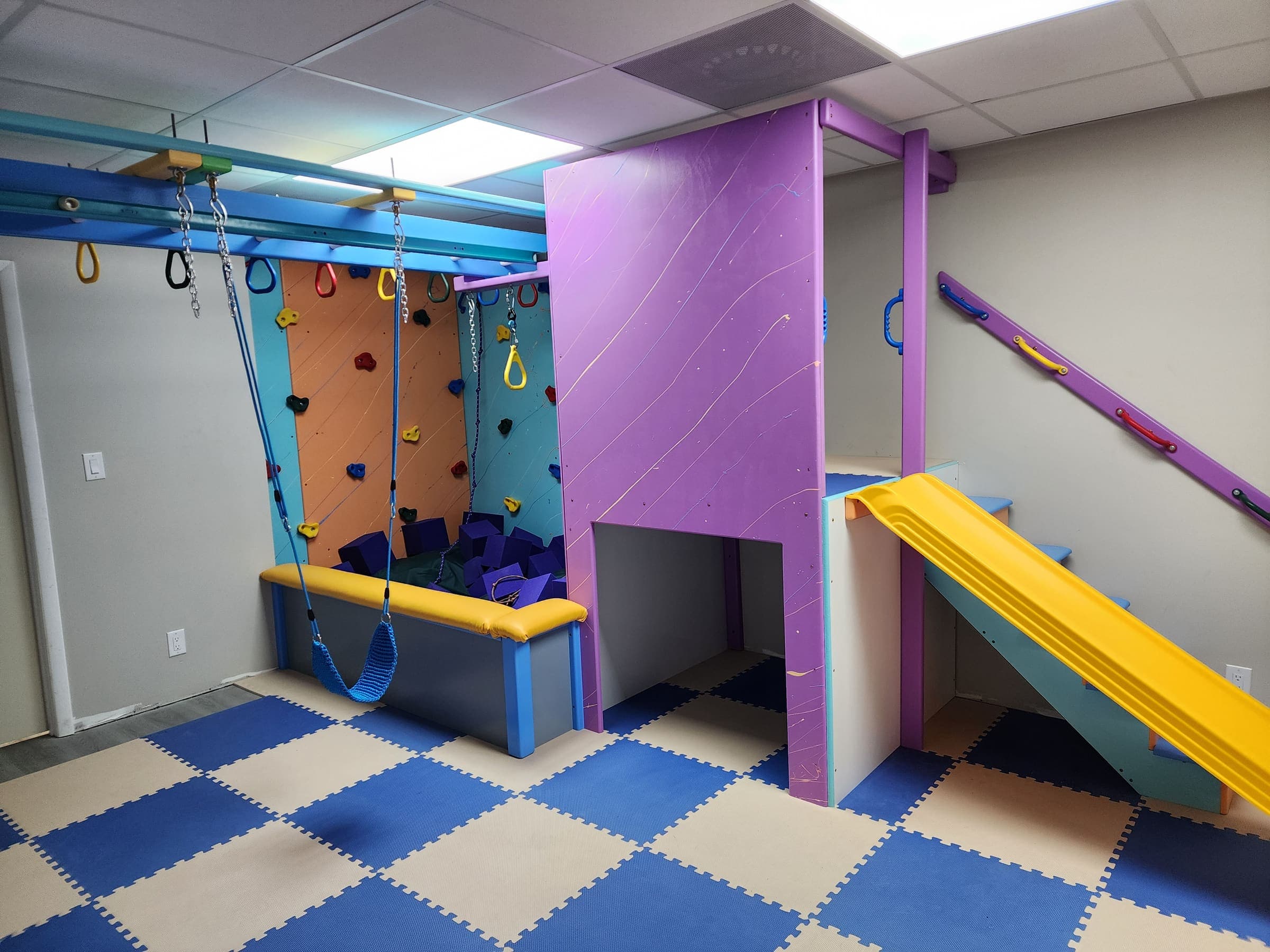 Completed Spectrum clinic sensory gym with purple playhouse, yellow slide, foam pit, blue swing, and climbing walls